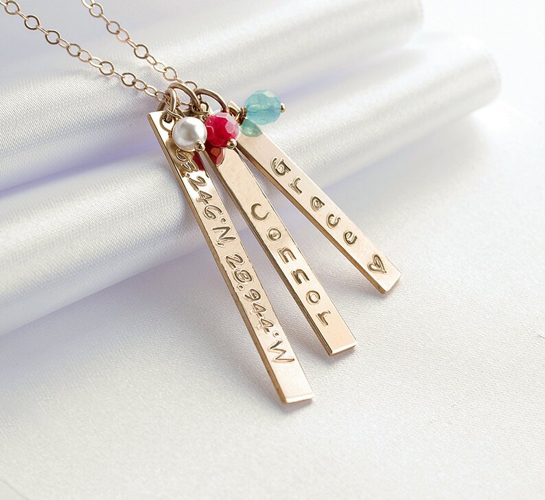 May include: Gold necklace with three bar pendants. The pendants are engraved with the words "Joescol", "Bless", and "32.46&deg;N 28.9444&deg;W". The necklace also features a small pearl and a red and blue gemstone.