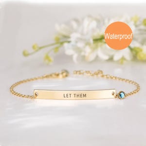May include: A gold-toned chain bracelet featuring a central curved metal bar engraved with the text LET THEM. The bracelet includes a small, round, blue gemstone accent set in a gold-toned bezel on one side of the bar and a clasp closure.