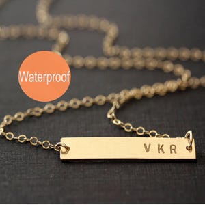 May include: A gold-toned bar necklace featuring a delicate chain and a rectangular pendant engraved with the letters V K R. An orange circular graphic with the white text Waterproof is positioned in the upper left area of the image.