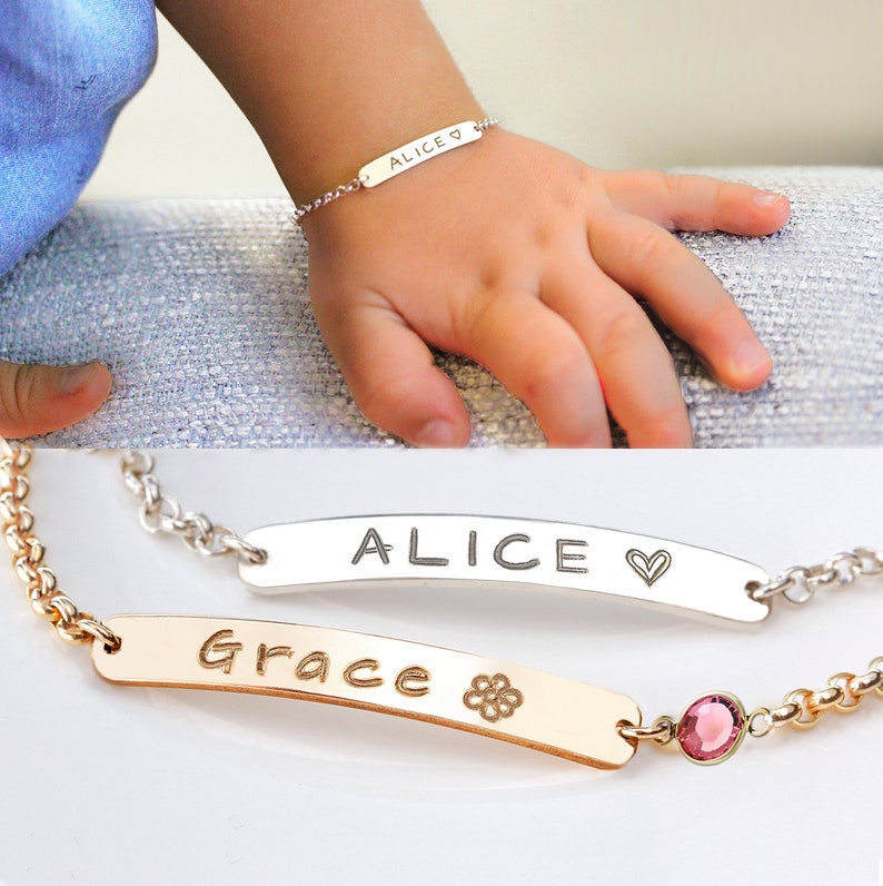 May include: Two personalised nameplate bracelets. The top bracelet is silver with the name "Alice" and a heart charm. The bottom bracelet is gold with the name "Grace" and a flower charm. The bracelet also has a pink gemstone.