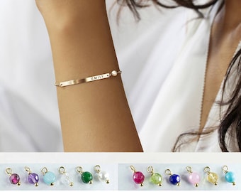 May include: Gold bracelet with a personalized nameplate that reads "EMILY" and a small pearl charm. The bracelet is shown on a person's wrist. The image also shows a variety of gemstone charms in different colors, including red, purple, blue, green, and white.