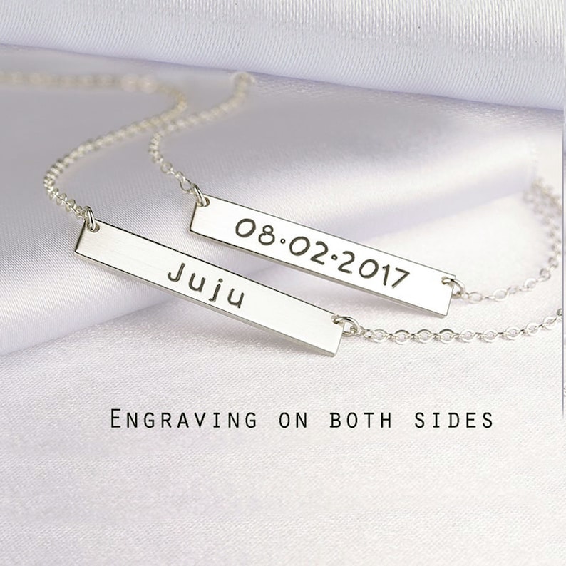 May include: Two silver-toned bar necklaces featuring delicate chains. One bar is engraved with the name Juju, and the other bar displays the date 08.02.2017. The text Engraving on both sides is visible below the jewelry pieces.