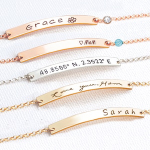 May include: Four gold bar bracelets with different engravings. The top bracelet has the word "Grace" and a flower symbol. The second bracelet has the words "Mom" and a heart symbol. The third bracelet has the coordinates "48.8566&deg; N, 2.3522&deg; E". The bottom bracelet has the words "Love you, Mom" in cursive.