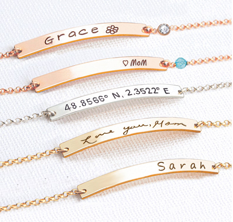 May include: Four gold bar bracelets with different engravings. The top bracelet has the word "Grace" and a flower symbol. The second bracelet has the words "Mom" and a heart symbol. The third bracelet has the coordinates "48.8566&deg; N, 2.3522&deg; E". The bottom bracelet has the words "Love you, Mom" in cursive.