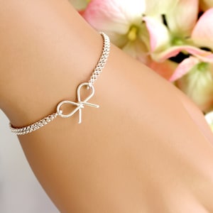May include: A silver chain bracelet with a delicate bow charm. The bracelet is on a person's wrist.