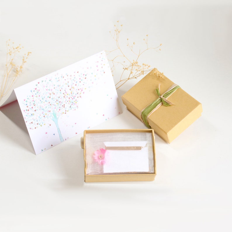 May include: A small gift box with a brown paper exterior and a green ribbon tied in a bow. The box is open and contains a small, white card with the word "Mom" written on it. A pink flower is placed on the card. The box is sitting on a white surface.