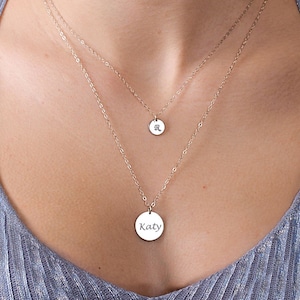 May include: Two silver necklaces with round pendants. The top pendant has the letter "R" engraved on it. The bottom pendant has the name "Katy" engraved on it.