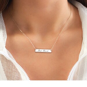 May include: A gold-toned chain necklace featuring a horizontal rectangular bar pendant. The pendant is engraved with the text "Best Friends" in a cursive script font. The necklace is worn around the neck, showcasing the delicate metal link chain.