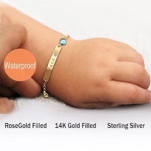 May include: A gold-toned identification bracelet featuring a rectangular metal plate engraved with the name Grace and a small flower icon, accented by a single round blue gemstone and a delicate chain. Text includes Waterproof, RoseGold Filled, 14K Gold Filled, and Sterling Silver.