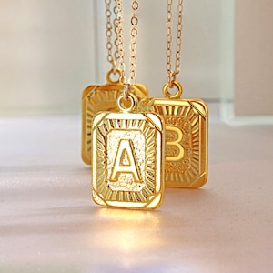 May include: Three gold necklaces with square pendants featuring the letters A, B, and C. The pendants have a textured, sunburst design.