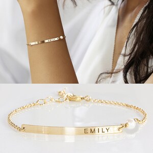 May include: A gold-toned metal chain bracelet featuring a thin, horizontal rectangular bar engraved with the name EMILY in capital letters. The bracelet includes a small, round white pearl accent attached to one side of the bar and a lobster clasp.