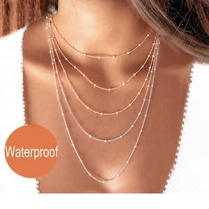 May include: A layered necklace set featuring multiple delicate chains with small, evenly spaced beads. The set includes both gold-toned and silver-toned chains of varying lengths, creating a cascading effect. An orange circle with the text Waterproof.