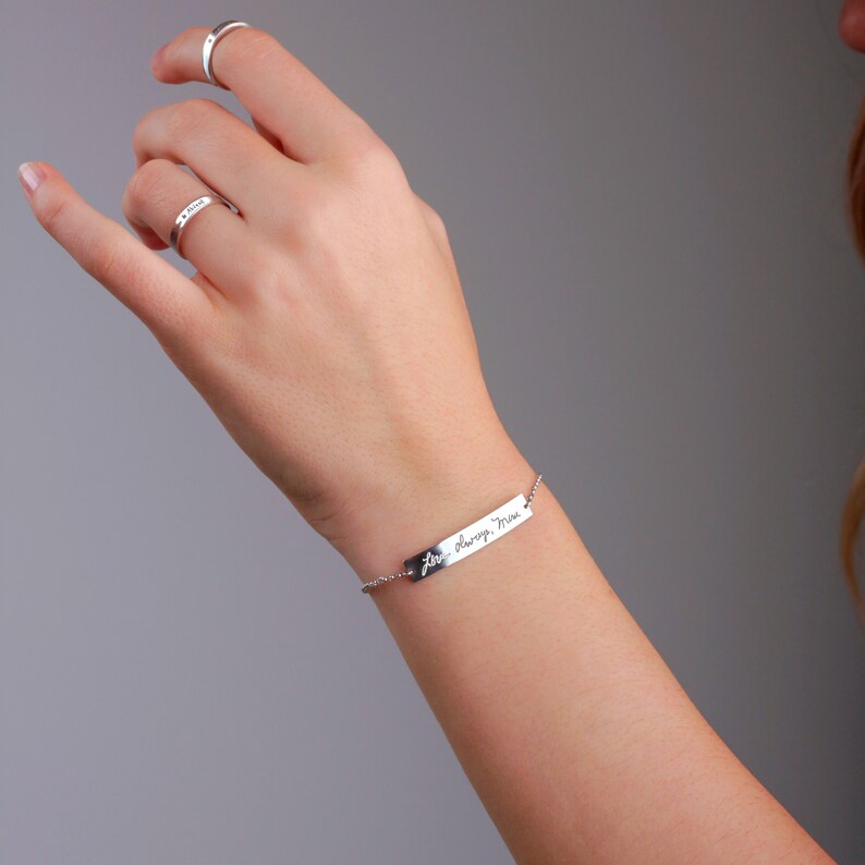 May include: A silver bar bracelet featuring the engraved cursive text Love always, Mom, attached to a delicate chain. Two matching silver rings are worn on the fingers, one plain and one engraved with the name Mom in a simple script font.