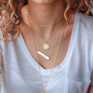 May include: A woman wearing a white t-shirt with a layered gold necklace set. The necklace features a coin pendant, a bar pendant with the word "Selah" inscribed, and a delicate chain.