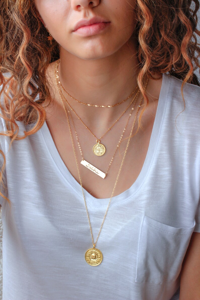 May include: A woman wearing a white t-shirt with a layered gold necklace set. The necklace features a coin pendant, a bar pendant with the word "Selah" inscribed, and a delicate chain.