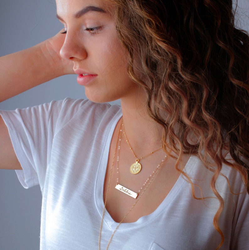 May include: A woman wearing a white t-shirt with a gold necklace featuring a delicate bar pendant with the word "Debbie" and a round coin pendant.