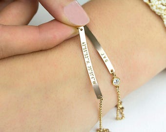 May include: Two gold-toned metal bar bracelets with delicate chains. One bar is engraved with the coordinates 42°55'53" N, 75°51'4" W, and the other bar is engraved with the letters J V G. A small, round, clear stone is set on one chain.