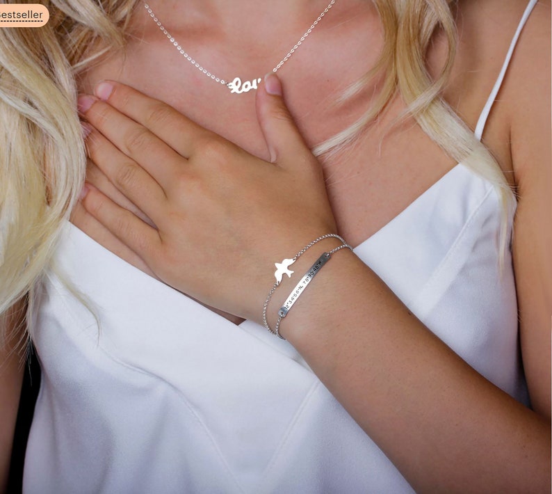 May include: A silver-toned necklace featuring the word love in cursive script and a double-strand silver-toned bracelet with a bird charm and a rectangular bar pendant engraved with coordinates, both crafted from polished metallic material.