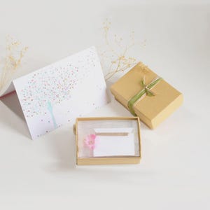 May include: A gold-toned bar necklace rests inside an open tan cardboard gift box with a pink flower accent. Beside it sits a closed tan gift box tied with a green ribbon and a white greeting card featuring a tree made of small colorful hearts.