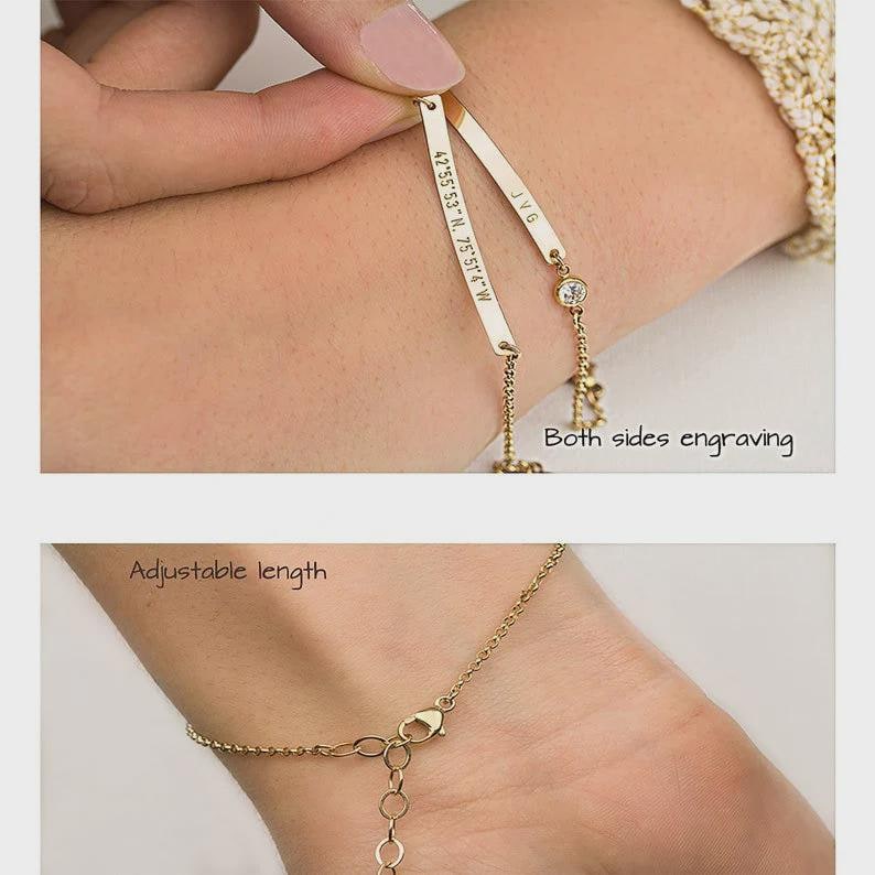 May include: A gold-toned bracelet featuring two thin, flat metal bars with engraved text, including coordinates and initials, alongside a small round clear stone. The chain includes a lobster claw clasp and an adjustable length link section.