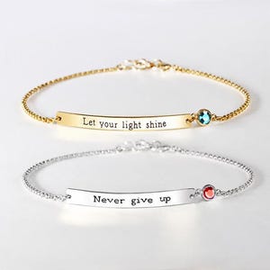 May include: Two delicate chain bracelets. The top gold-tone bracelet features a curved bar engraved with the text "Let your light shine" and a small blue crystal. The bottom silver-tone bracelet has a bar engraved with "Never give up" and a red crystal.