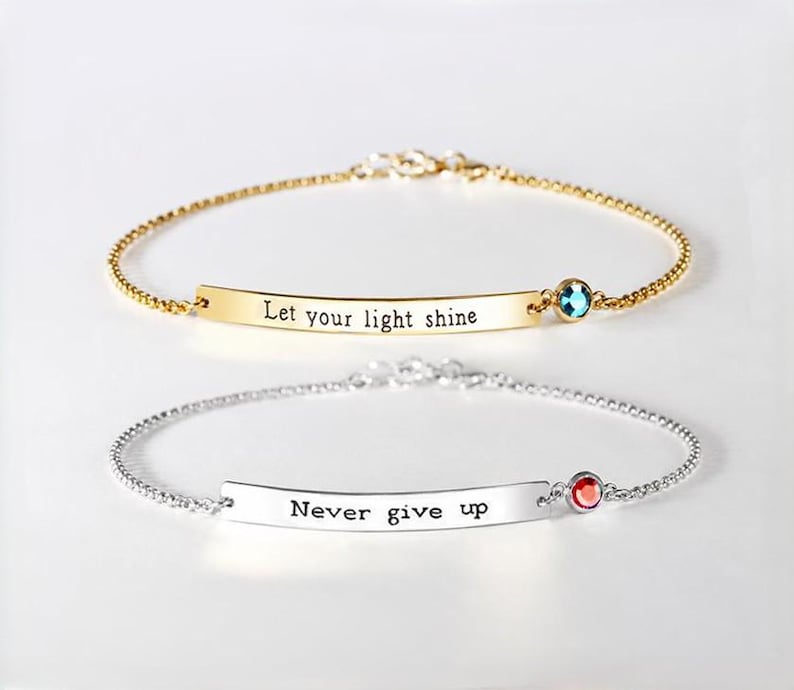 May include: Two delicate chain bracelets. The top gold-tone bracelet features a curved bar engraved with the text "Let your light shine" and a small blue crystal. The bottom silver-tone bracelet has a bar engraved with "Never give up" and a red crystal.