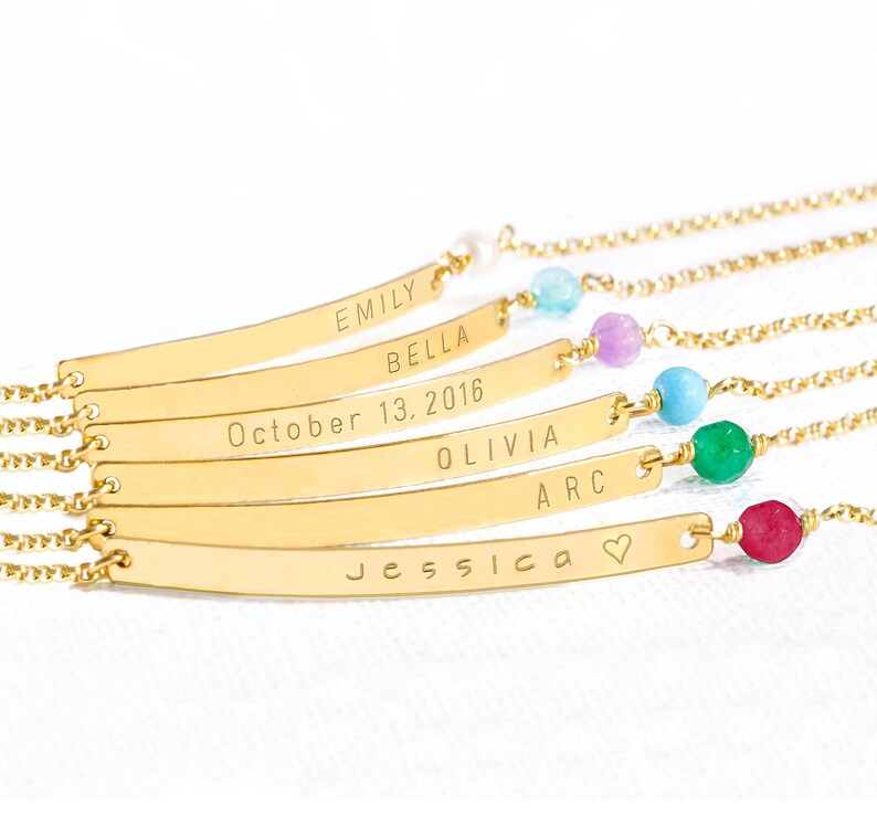 May include: Six gold bar bracelets with different names and dates engraved on them. Each bracelet has a small gemstone bead in a different color: light blue, purple, turquoise, green, and red.