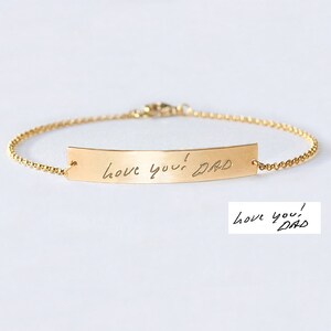 May include: A gold-toned metal bracelet featuring a thin chain and a curved rectangular bar. The bar is engraved with the handwritten text "love you! DAD" in black script. A separate white rectangular insert displays the same handwritten text.