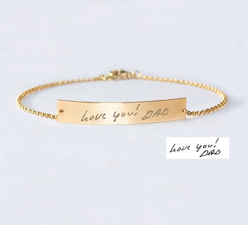 May include: A gold-toned metal bracelet featuring a thin chain and a curved rectangular bar. The bar is engraved with the handwritten text "love you! DAD" in black script. A separate white rectangular insert displays the same handwritten text.