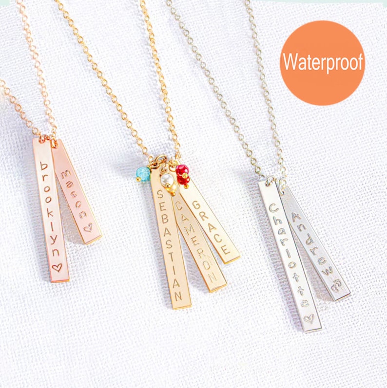 Op de afbeelding: Drie gouden bar kettingen met gepersonaliseerde namen, "Brooklyn", "Mason", "Sebastian", "Cameron" en "Grace", en een zilveren bar ketting met gepersonaliseerde namen, "Andrew" en "Charlotte". De kettingen zijn versierd met kleine kralen in turquoise, wit en rood. De afbeelding bevat het woord "Waterproof".