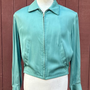 May include: A teal jacket with a collared neckline and a full-length zip. The jacket features long sleeves, two front pockets, and a cinched waist. It is displayed on a mannequin against a reddish-brown background.