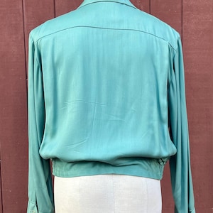 May include: A teal long-sleeved shirt with a collared neckline and buttoned cuffs. The shirt features a gathered waist and a relaxed fit. The fabric has a subtle sheen. The back of the shirt is visible.