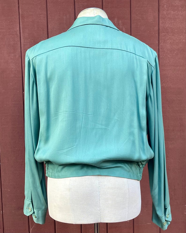 May include: A teal long-sleeved shirt with a collared neckline and buttoned cuffs. The shirt features a gathered waist and a relaxed fit. The fabric has a subtle sheen. The back of the shirt is visible.