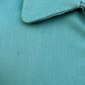May include: Close-up of teal fabric, possibly part of a shirt or garment. The collar is visible, with white stitching along the edges. The fabric has a subtle texture, and the overall appearance is clean and simple.