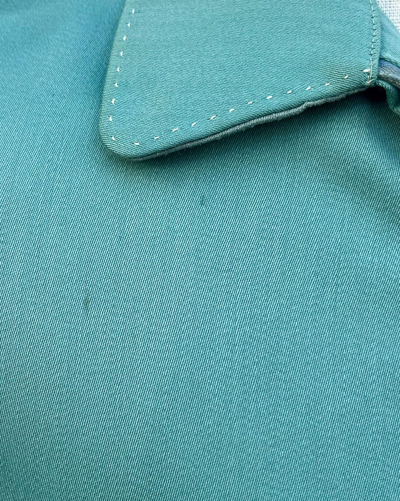May include: Close-up of teal fabric, possibly part of a shirt or garment. The collar is visible, with white stitching along the edges. The fabric has a subtle texture, and the overall appearance is clean and simple.