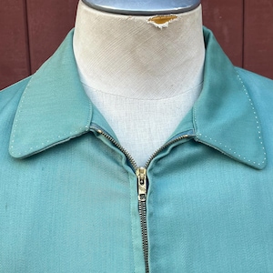May include: A teal jacket with a full brass zip and a classic collar, displayed on a mannequin. The fabric looks lightweight, suitable for warmer weather. Stitching is visible along the collar and seams. The jacket is a vintage style.