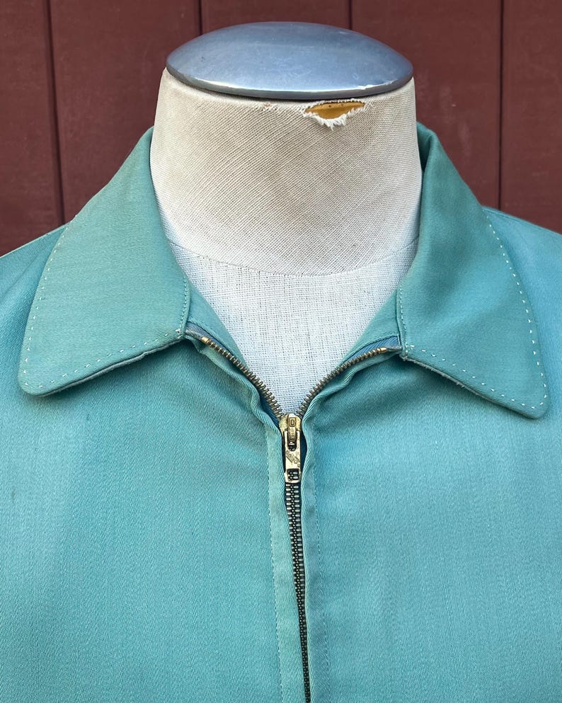 May include: A teal jacket with a full brass zip and a classic collar, displayed on a mannequin. The fabric looks lightweight, suitable for warmer weather. Stitching is visible along the collar and seams. The jacket is a vintage style.