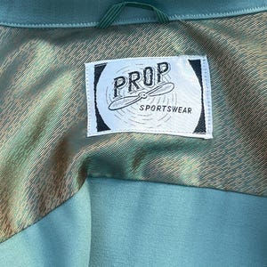 May include: Detail of a teal garment's lining, featuring a label with "PROP SPORTSWEAR" and a propeller design. The lining has a metallic, textured finish, contrasting with the teal exterior fabric.