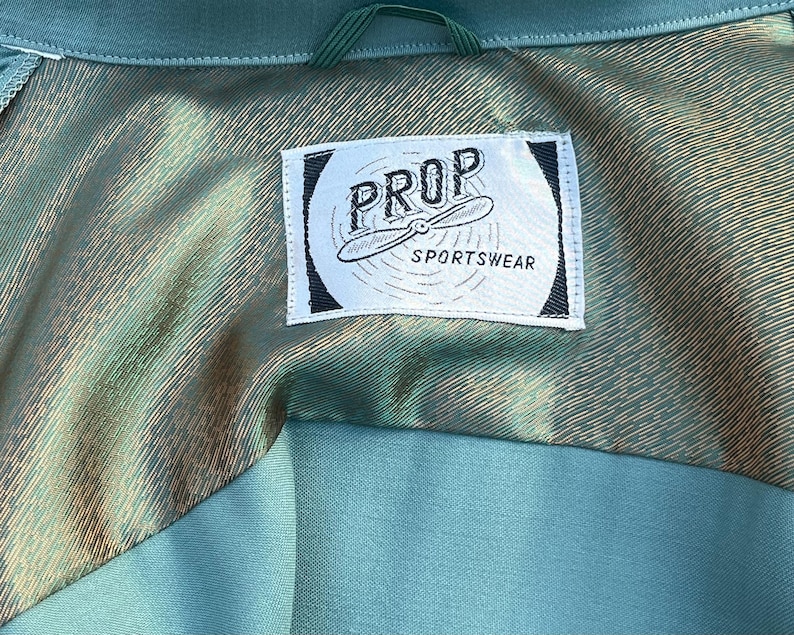 May include: Detail of a teal garment's lining, featuring a label with "PROP SPORTSWEAR" and a propeller design. The lining has a metallic, textured finish, contrasting with the teal exterior fabric.