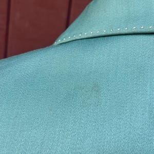 May include: Close-up of a teal blazer collar. The collar features white stitching along the edges. The fabric has a subtle texture, and a small dark mark is visible on the material.