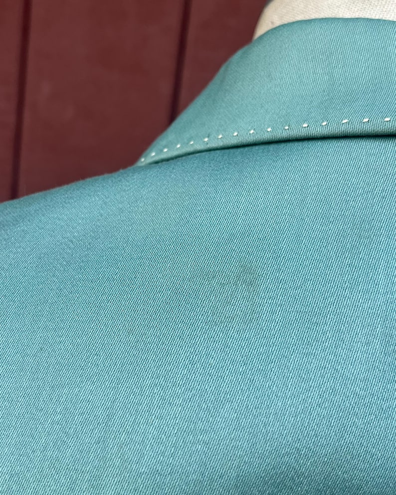May include: Close-up of a teal blazer collar. The collar features white stitching along the edges. The fabric has a subtle texture, and a small dark mark is visible on the material.