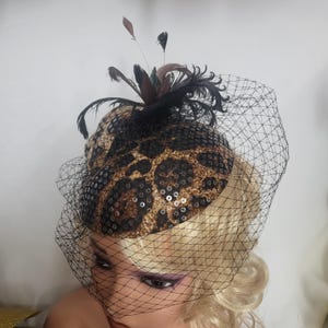 May include: A fascinator hat featuring a leopard print pattern with black sequin embellishments. The headpiece is adorned with dark brown and black feathers and includes a black netting veil that drapes over the front of the headpiece.