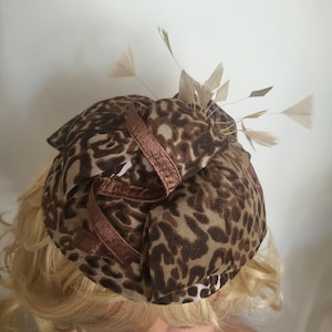 May include: A fascinator hat featuring a brown and tan leopard print pattern on a textured fabric base. The headpiece is adorned with a dark brown ribbon trim and several light beige feathers extending outward from the top center section.