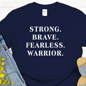 May include: A navy blue t-shirt with the words "STRONG. BRAVE. FEARLESS. WARRIOR." in white. The shirt is paired with light blue jeans, yellow flowers, brown sunglasses, and tan sandals with blue and white stripes.
