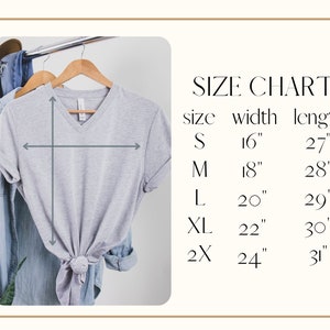 May include: A light gray V-neck t-shirt hanging on a wooden hanger, with a size chart to the right. The size chart lists sizes S to 2X, with corresponding width and length measurements in inches. The shirt is styled with a knot at the bottom.