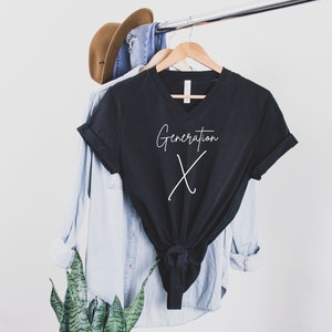May include: A black V-neck t-shirt with the words "Generation X" in white script. The shirt is hanging on a wooden hanger, alongside a light blue button-down shirt, a pair of blue jeans, and a brown hat.