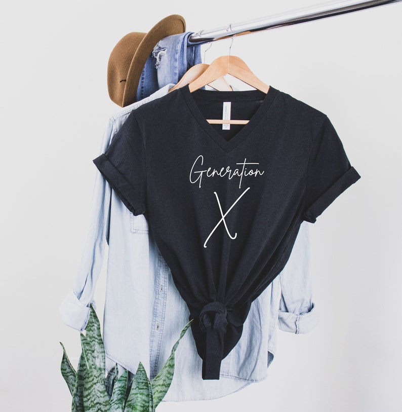May include: A black V-neck t-shirt with the words "Generation X" in white script. The shirt is hanging on a wooden hanger, alongside a light blue button-down shirt, a pair of blue jeans, and a brown hat.