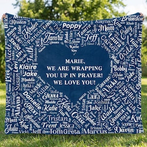 May include: A navy blue blanket with a heart-shaped design in the center, surrounded by names in white. The text in the center reads, "MARIE, WE ARE WRAPPING YOU UP IN PRAYER! WE LOVE YOU!" A personalized gift.
