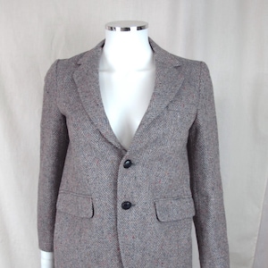 May include: A gray tweed blazer with a herringbone pattern. The jacket has two front pockets and two black buttons. The blazer is displayed on a mannequin with blue jeans.