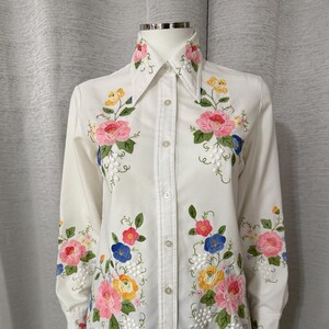 May include: A long-sleeved white button-down shirt featuring colorful floral embroidery. The design includes pink, yellow, and blue flowers with green leaves and small white beaded accents scattered across the front, collar, and sleeves.